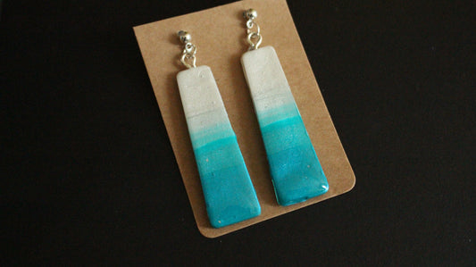 A pair of handcrafted rectangular earrings with a shimmering ombre design, fading from white at the top to aqua blue at the bottom, attached to silver ball studs and displayed on a brown card.