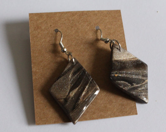 A pair of handcrafted diamond-shaped earrings with a marbled swirl pattern in black, gold, and bronze tones, finished with a subtle shimmer, displayed on a brown card.